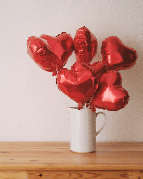 a red heart shaped balloon bouquet displayed in a white decorative jug